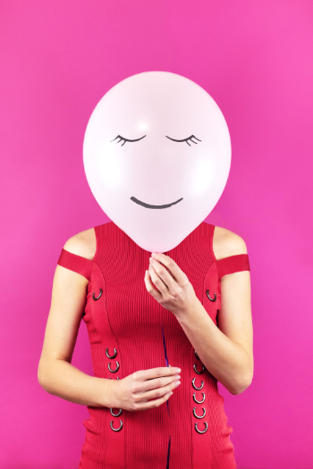 Eine Frau im roten Kleid hält sich einen rosa Ballon vors Gesicht, auf den ein lächelndes Gesicht mit geschlossenen Augen gemalt ist. Denn der core unseres Körpers lässt sich mit einem Luftballon vergleichen, deshalb eignen sich SItups nicht als Übungen nach der Schwangerschaft